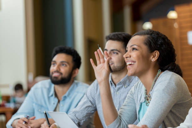 Confident mid adult college student raises her hand to ask or answer question while attending a college class.