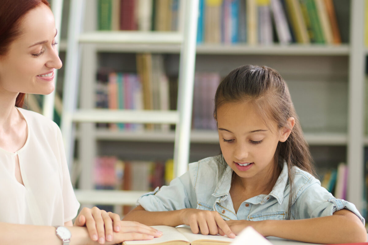 Private lesson. Smiling attentive young long-haired woman attentively watching listening school-age girl reading book sitting at table in library